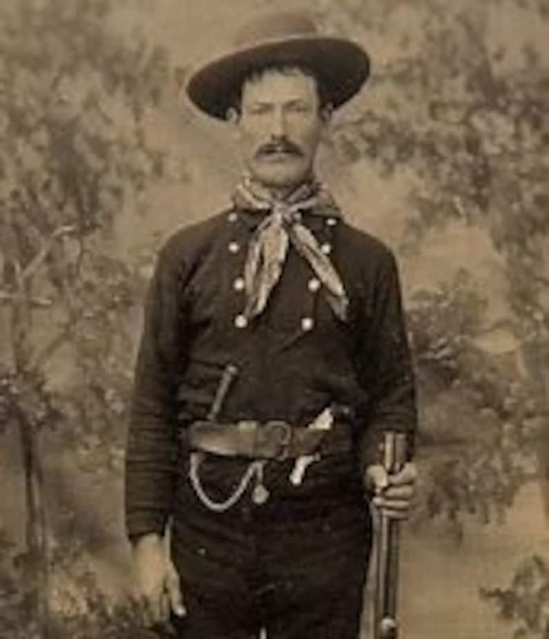 May include: A man wearing a dark blue shirt with a light blue neckerchief, a brown leather belt with a silver buckle, and a brown cowboy hat. He is holding a rifle in his right hand.