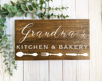 May include: A rectangular dark brown wooden sign featuring the white text Grandma's KITCHEN & BAKERY. Below the text, there are white graphic silhouettes of a spoon, a fork, and a knife arranged horizontally across the bottom of the wooden board.