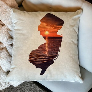 May include: A throw pillow with a silhouette of the state of New Jersey filled with a sunset over the ocean. The pillow is beige and has a linen texture.