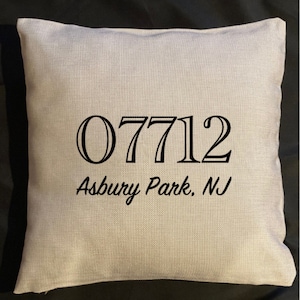 May include: A square, beige throw pillow with the black text "07712 Asbury Park, NJ" printed on the front. The pillow has a textured appearance and is likely made of linen or a similar fabric. The background is black.