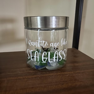 May include: A clear glass jar with a silver lid filled with sea glass. The jar has a white label with the text "I want to age like sea glass".