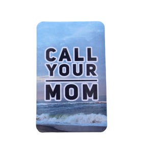 May include: A rectangular sticker with the text "CALL YOUR MOM" in bold, white letters with a black outline. The background features a scenic view of the ocean and sky.