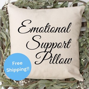 May include: A white pillow with black cursive text that reads "Emotional Support Pillow". The pillow is sitting in a woven basket with green leafy branches.