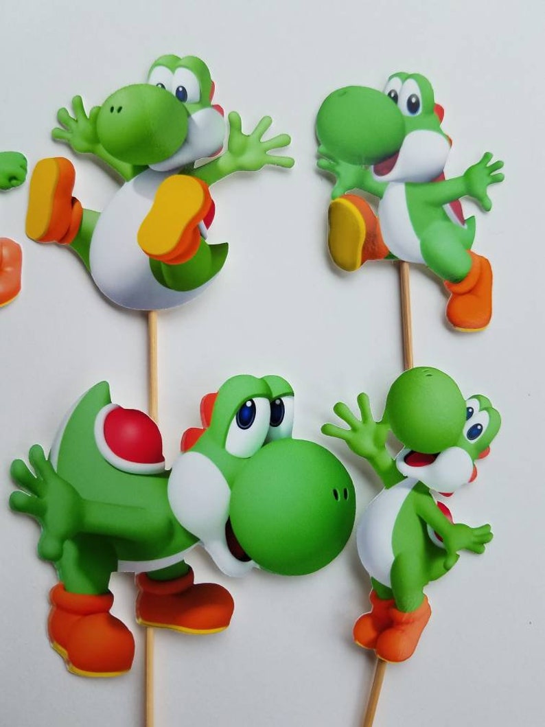 May include: Four green Yoshi cake toppers with different poses. Each topper is made of cardboard and has a stick attached to the back.