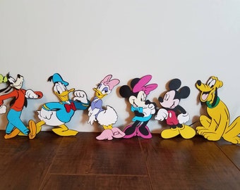 May include: A set of seven colorful cartoon character cutouts featuring Mickey Mouse, Minnie Mouse, Donald Duck, Daisy Duck, Goofy, Pluto, and a green butterfly. The characters are all standing in a row on a wooden floor.