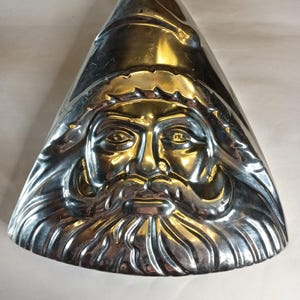 May include: A silver-colored, triangular mold in the shape of Santa Claus's face. The mold features a detailed depiction of Santa's face, including his hat, beard, and facial features. The mold is made of a shiny metal and is likely used for baking or decorative purposes.
