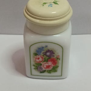 May include: A small, white ceramic jar with a lid. The lid and the jar's base are a creamy off-white color. The jar features a floral design on the front, with pink, purple, and blue flowers. The lid has a small floral design.