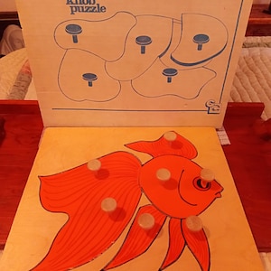 May include: A vintage wooden knob puzzle featuring a goldfish design. The puzzle is orange and black with wooden knobs for easy handling. The top board reads "knob puzzle" and "CHILDCRAFT".