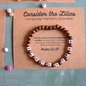 Puede incluir: Una pulsera de cuentas de madera con cuentas de letras blancas que deletrean "FOOTLIT MOM" se muestra en una tarjeta marrón. La tarjeta dice "Consider the Lilies Scripture memory bracelet" con un versículo bíblico. Se ven otras pulseras y cuentas.