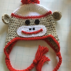 May include: A crocheted monkey hat with a red pom pom and a long red braided strap with a tassel. The hat is brown, white, and red.