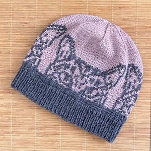 Puede incluir: Un gorro de punto en tonos rosa empolvado y gris carbón. El gorro presenta un diseño estampado en la parte superior, con una banda gris carbón lisa en la parte inferior. El gorro está hecho de un material suave y cálido.