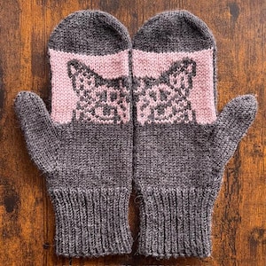 Könnte beinhalten: Ein Paar graue Strickfäustlinge mit einem rosa Katzendesign auf dem Handrücken. Die Fäustlinge sind aus weichem, strukturiertem Garn gefertigt und haben gerippte Bündchen. Das Katzendesign ist detailliert.