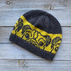Pode incluir: Um gorro de malha cinza com um padrão geométrico amarelo. O chapéu apresenta uma bainha canelada e uma faixa amarela contrastante com um design repetido. O gorro é feito de um material macio e quente, ideal para o frio.