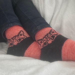 May include: A pair of knitted socks with a black and coral color scheme. The socks feature a black band around the ankle with a decorative pattern, and the toe and heel are coral. The socks are worn with blue jeans.
