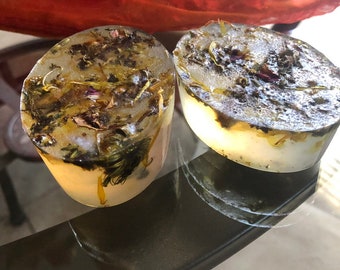 May include: Two clear, round soaps with dried flowers and herbs embedded inside. The soaps are on a glass surface.