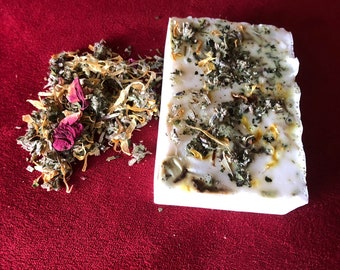 May include: A white bar of soap with dried herbs and flowers embedded in it. The soap is sitting on a red surface.