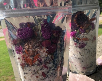 May include: Two silver foil pouches filled with white bath salts and dried flowers. The flowers are purple, pink, and red. The pouches are sitting on a surface with pink and white rocks.