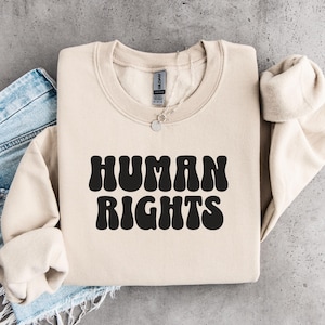 May include: A beige sweatshirt with the words "HUMAN RIGHTS" in bold, black, retro-style lettering. The sweatshirt is folded with the sleeves rolled up. A pair of blue jeans is visible on the left side of the image.