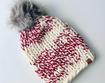 May include: A chunky knit beanie in cream and red stripes, topped with a gray faux fur pom. The hat features a small brown leather tag. This winter hat is a cozy accessory.
