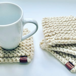 May include: Off-white, hand-knitted coasters with a chunky texture. A white mug rests on one coaster, with a stack of three more coasters nearby. Each coaster has a small, brown leather tag that reads "Livnloop".