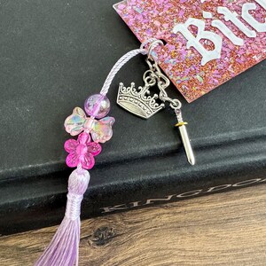 Pu&ograve; includere: Un segnalibro fatto a mano con un nappa viola, perline iridescenti e accenti floreali rosa. Presenta un ciondolo a forma di corona e spada argentata e un'etichetta rosa glitterata con la parola "Bitch" in lettere bianche.