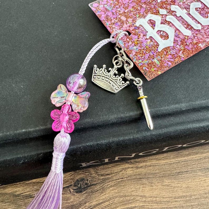Pu&ograve; includere: Un segnalibro fatto a mano con un nappa viola, perline iridescenti e accenti floreali rosa. Presenta un ciondolo a forma di corona e spada argentata e un'etichetta rosa glitterata con la parola "Bitch" in lettere bianche.