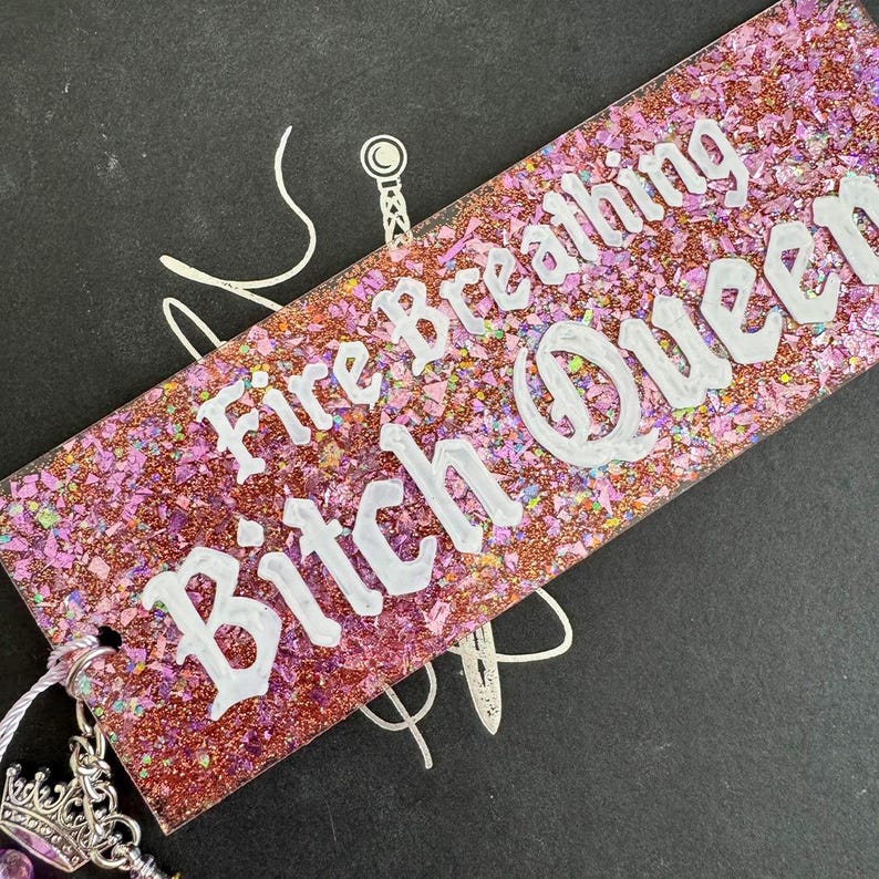 Pu&ograve; includere: Un segnalibro rettangolare e glitterato con la scritta "Fire Breathing Bitch Queen" in bianco. Il segnalibro ha un design glitterato rosa e viola. Un ciondolo argentato con una corona e perline viola pende dalla parte superiore.