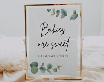 May include: A gold-framed sign with a white background and green eucalyptus leaves. The sign reads "Babies are sweet" and "Please take a treat".