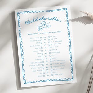 Peut inclure: Une feuille de papier blanche avec une bordure bleue festonnée et le texte "Would she rather". Le papier contient une liste de questions pour un jeu de fête prénuptiale. Le texte est écrit dans une police manuscrite.