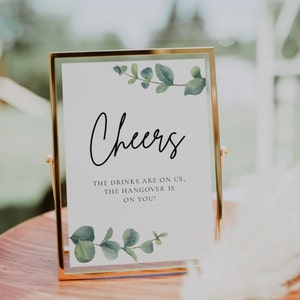 Puede incluir: Un cartel con marco dorado, fondo blanco y hojas de eucalipto verdes en la parte superior e inferior. El cartel dice "Cheers" en letras cursivas negras y "The drinks are on us, the hangover is on you!"