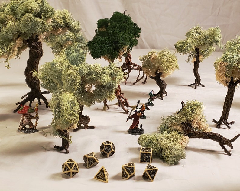 May include: A tabletop scene with miniature figures and dice, surrounded by artificial moss trees. The scene features a variety of trees, including a large green tree, a smaller green tree, and several white moss trees. The miniature figures are painted in various colors and are standing on a brown surface. The dice are black and white and are scattered around the figures.