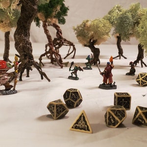 May include: A miniature tabletop game scene with a variety of figures, including a mushroom-shaped creature, a goblin, and a warrior. The scene is set in a forest with trees and moss. There are also several dice, including a black and gold d20, a black and gold d12, a black and gold d8, a black and gold d6, and a black and gold d4.
