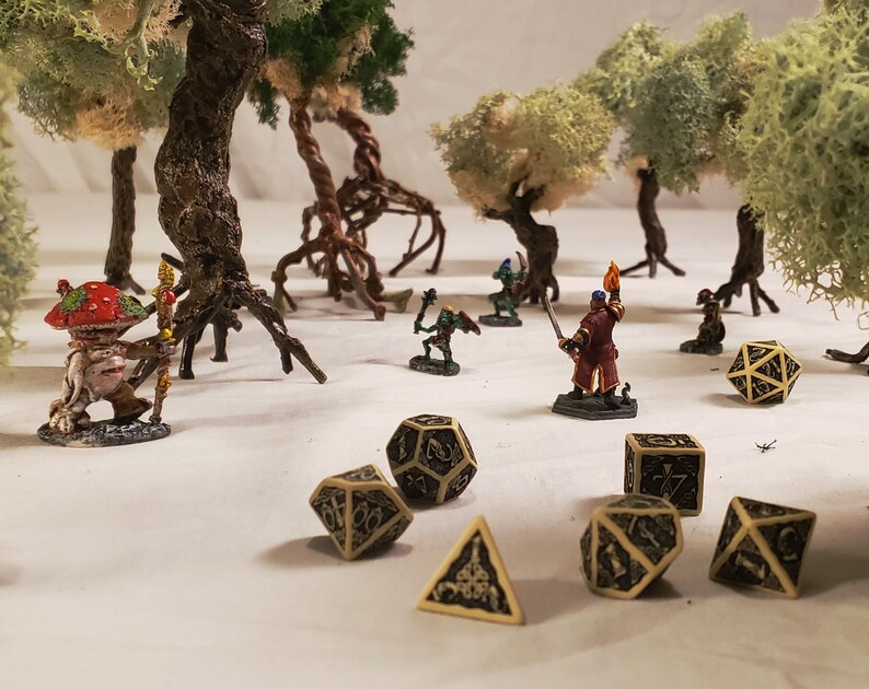 May include: A miniature tabletop game scene with a variety of figures, including a mushroom-shaped creature, a goblin, and a warrior. The scene is set in a forest with trees and moss. There are also several dice, including a black and gold d20, a black and gold d12, a black and gold d8, a black and gold d6, and a black and gold d4.