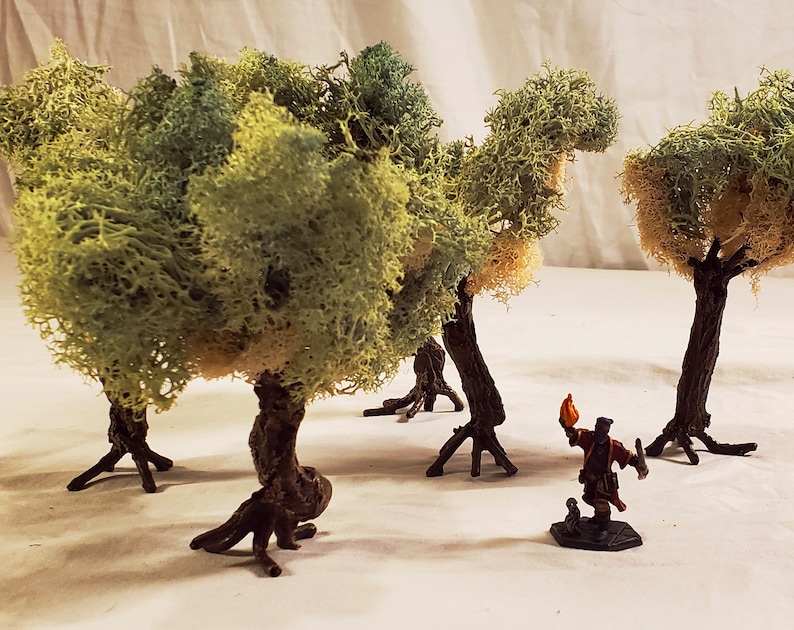 May include: Four miniature trees made of green and brown moss with a small figurine of a person holding a torch standing in front of them.