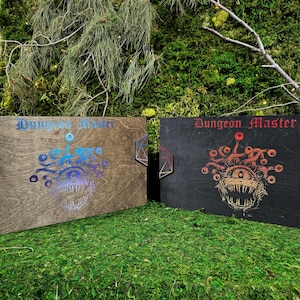 Puede incluir: Dos cajas de madera con el texto "Dungeon Master". Una caja es marrón con un diseño de monstruo azul y morado, la otra negra con un diseño de monstruo rojo y naranja. Se exhiben sobre musgo verde.