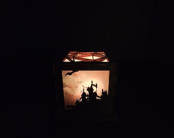 May include: A wooden lantern with a silhouette of a castle and bats against a full moon. The lantern is lit from within, casting a warm glow.