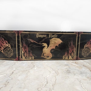 May include: A three-piece wooden game board with a black stained finish. The center panel features a gold dragon with flames and the text "Dungeon Master". The side panels feature a gold 20-sided die with flames.