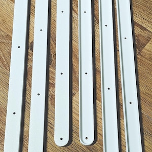 May include: Six white, elongated, rectangular pieces with rounded ends and evenly spaced holes. The pieces are arranged on a wooden surface. The pieces appear to be made of metal or a similar material.