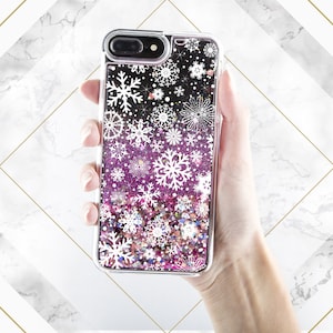 May include: A clear phone case with a glittery pink and white design featuring snowflakes. The case is made of a clear plastic material and has a raised lip around the edges to protect the screen.