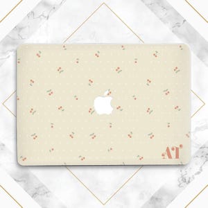 Op de afbeelding: Een crèmekleurige laptophoes met een herhaald patroon van kleine rode kersen met groene blaadjes en witte stippen. Het Apple-logo is zichtbaar en de initialen "AT" staan in de rechter benedenhoek.