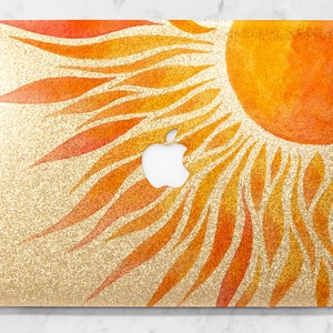 May include: A gold glitter laptop skin with a watercolor sun design in shades of orange and yellow.