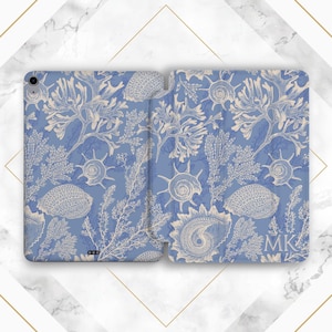 May include: A blue tablet case featuring a detailed cream-colored seashell and seaweed pattern. The case is open, revealing the interior design. The initials "MK" are visible in the lower right corner. The case is designed to protect a tablet.