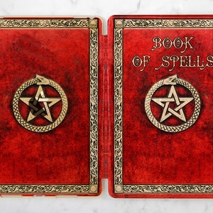May include: A red leather iPad case with a gold border and a pentagram with a snake wrapped around it. The text "BOOK OF SPELLS" is on the right side of the case.