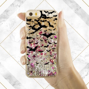 May include: A gold glitter phone case with a black and white spiderweb design and black bats. The case has a clear back and a glittery liquid inside.
