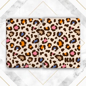 May include: A beige tablet case with a colorful leopard print design. The pattern includes brown, orange, pink, and blue spots. The initials "KM" are visible on the cover. The case is open, displaying the interior.