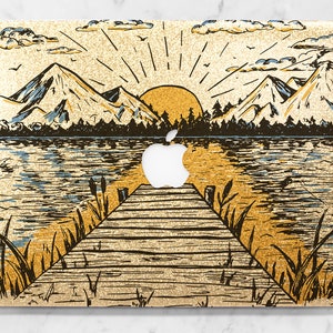May include: A gold glitter laptop skin with a hand-drawn illustration of a lake, mountains, and a sunrise. The illustration features a wooden dock extending into the water.