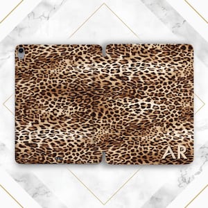 May include: A leopard print tablet case, open to display the interior. The case is brown and tan with a repeating spotted pattern. Gold-colored initials "AR" are visible on the right side of the case. The case is on a white and gray marble background.