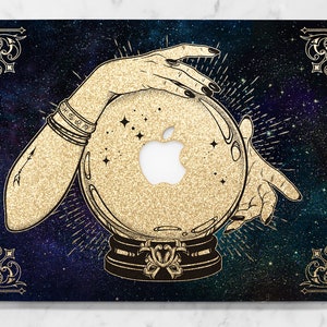 May include: A gold glitter laptop skin with a black background and a galaxy design. A hand with long nails holds a crystal ball with a gold glitter finish. The crystal ball has an Apple logo in the center.