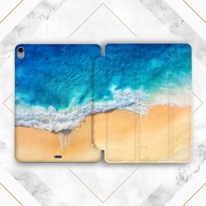 Op de afbeelding: Tablethoes met een levendig luchtfoto van een strand. Het ontwerp toont een helderblauwe oceaan die een zandstrand ontmoet met witte golven. De hoes is open en toont de binnenkant en de rand van het apparaat.