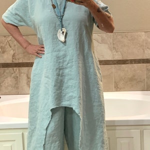 May include: A woman wearing a light blue linen dress with a shell pendant necklace and white sneakers. The dress has a high-low hemline and a relaxed fit. The woman is standing in a bathroom with a white bathtub and tiled walls.